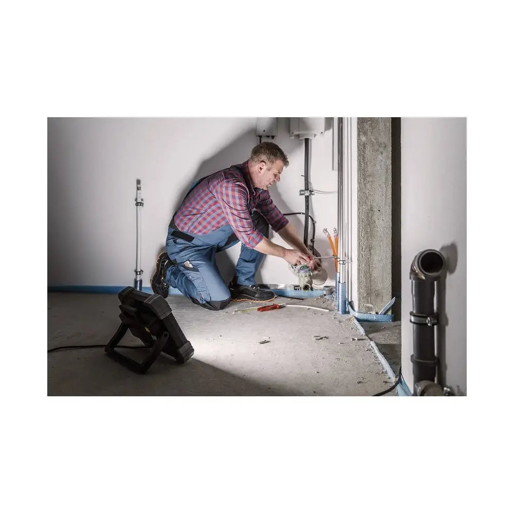 Arbeidslys LED DINORA 8061 lyser opp rørarbeid i hjørne, plassert på gulvet ved håndverker i arbeid.