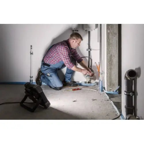 Arbeidslys LED DINORA 8061 i bruk under rørarbeid, montert i uferdig rom med synlige verktøy og rørdeler.