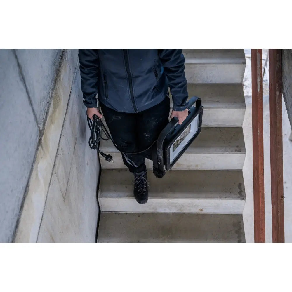 Arbeidslys LED JARO 20060 M bæres i hånd ned en betongtrapp, tydelig med bærehåndtak og ledning.