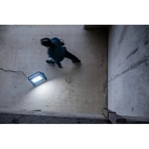 Arbeidslys LED JARO 20060 M, sett ovenfra mens det lyser opp betonggulv og en person passerer forbi.