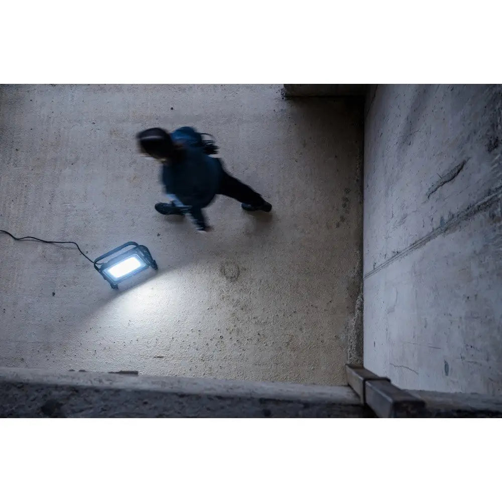 Arbeidslys LED JARO 20060 M, kraftig lampe på betonggulv som lyser mens person passerer forbi.