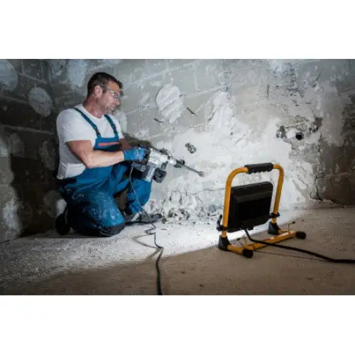 Arbeidslys LED JARO 7050 M med gul ramme lyser opp vegg mens håndverker borer, plassert på gulvet.