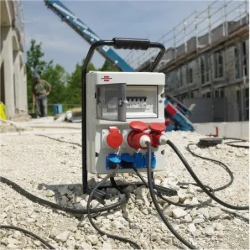 Bærbar byggsentral med røde og blå uttak, frontvisning på gruslagt byggeplass utendørs.