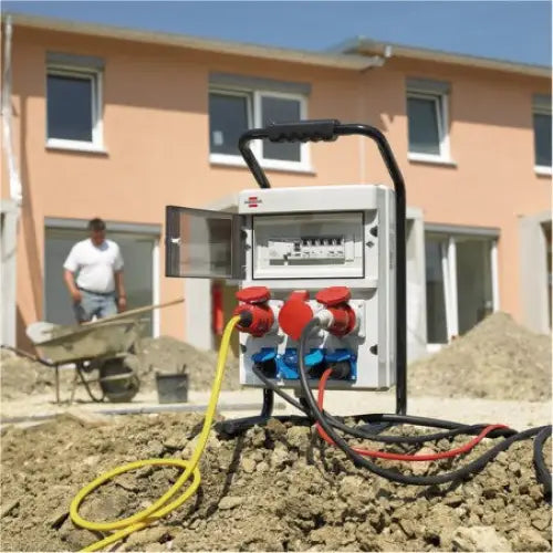 Bærbar byggsentral med fire tilkoblede kabler og åpen sikringsluke foran byggeplass med aktivitet.