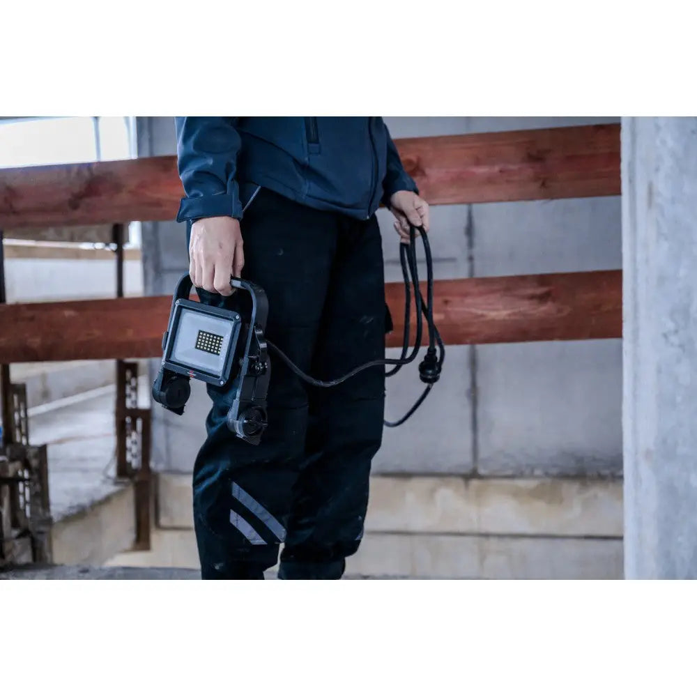 Bærbar LED-arbeidslampe holdt av person i arbeidsklær, sidevisning i industrielt verkstedmiljø.