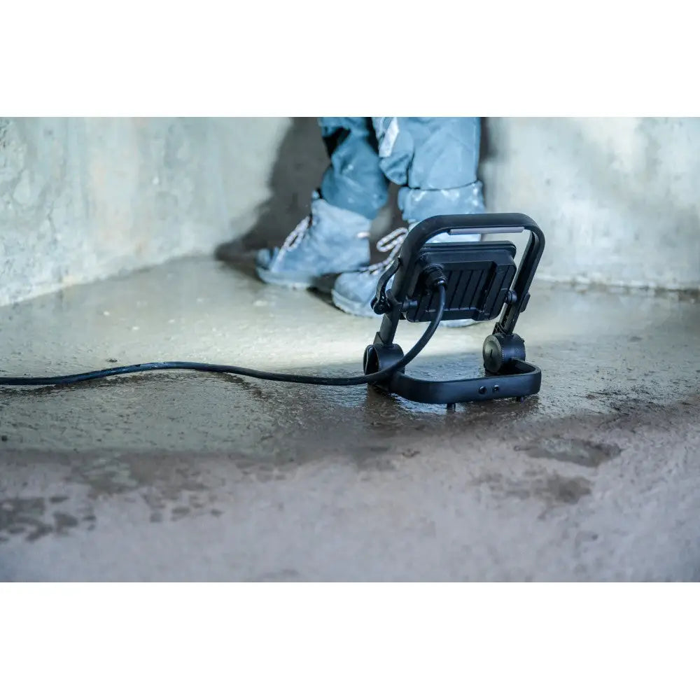 Bærbar LED arbeidslampe JARO, bakfra på vått underlag med person i arbeidsklær og støvler i bakgrunnen.