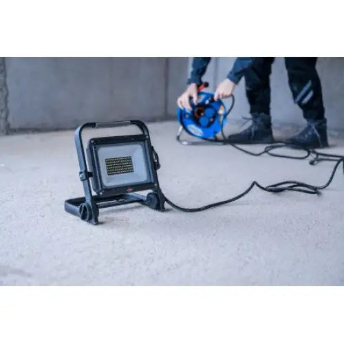 Bærbar LED-projektor JARO, svart lampe med stativ og strømledning, plassert på gulv i brukssituasjon.