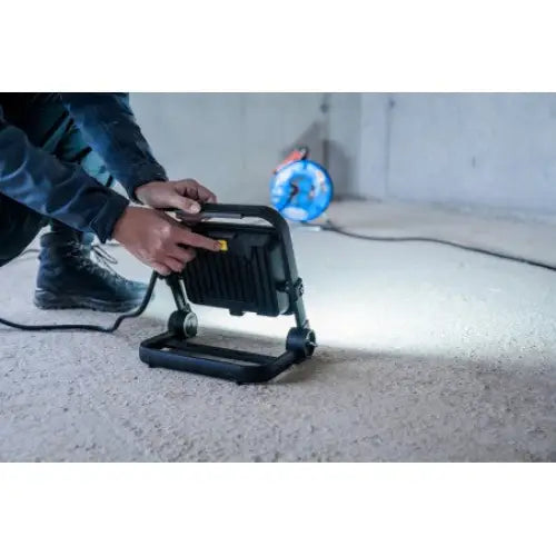 Bærbar LED-projektor JARO 7060, sort arbeidslampe justeres av en person på betonggulv med trommel i bakgrunnen.