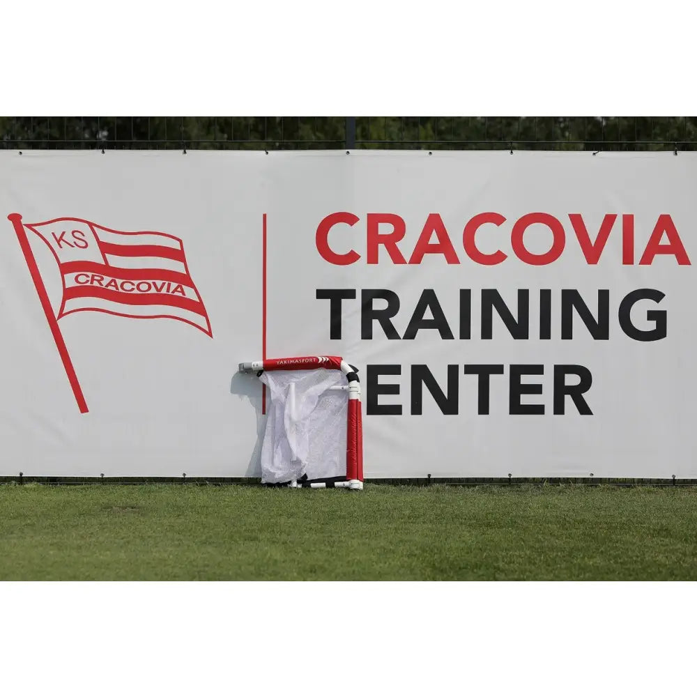 Sammenleggbart fotballmål med røde og hvite stenger, sett forfra ved skilt på gressbane inntil vegg.