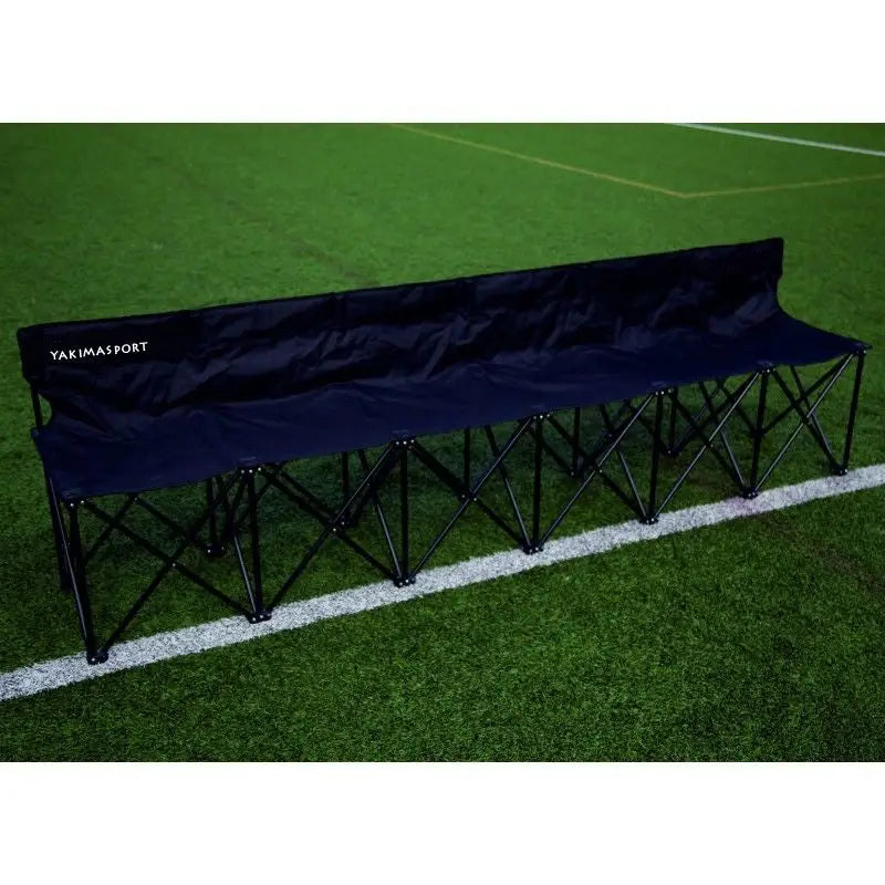 Sammenleggbar benk for seks personer i svart tekstil med ryggstøtte, sett skrått på kunstgressbane.