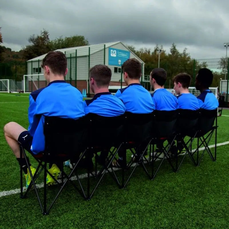 Sammenleggbar benk for seks personer, bakfra på kunstgressbane med fotballmål i bakgrunnen.