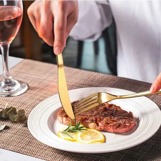 Gullfarget bestikk i bruk, kniv og gaffel skjærer biff med sitronskiver og rosmarin på hvit tallerken.