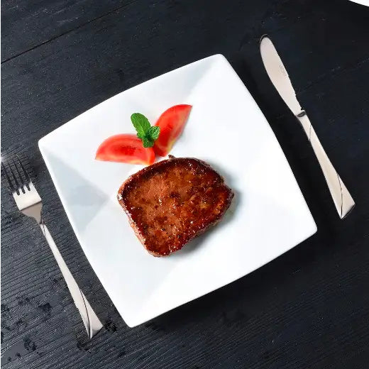 Bestikksett med kjøttbit på hvit tallerken, pyntet med tomat og mynte, sett ovenfra.