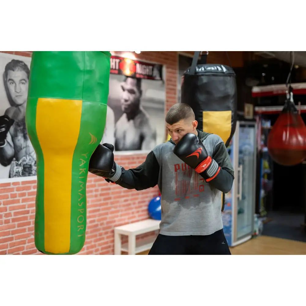 Boksehansker av ekte skinn PRO SPIDER i bruk under trening mot grønn og gul boksesekk i studio med rød murvegg.