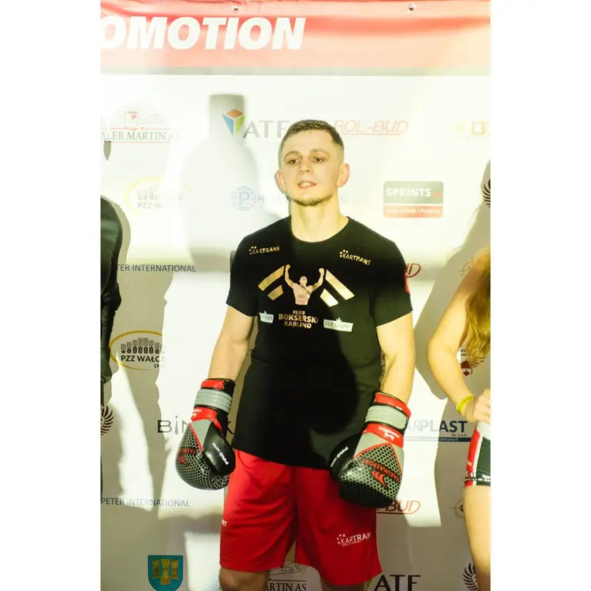 Boksehansker i ekte skinn PRO SPIDER, svart med røde detaljer, på mann i treningsantrekk foran reklamevegg.