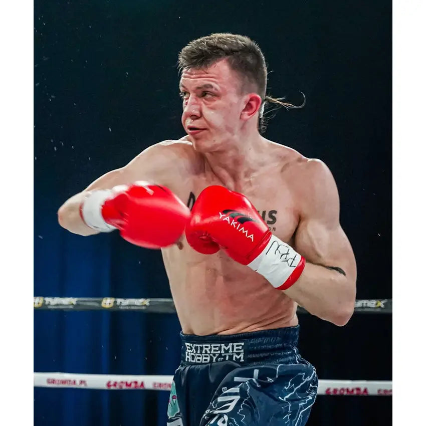 Boksehansker WOLF RØD, brukt av bokser i kampstilling med blå shorts og boksering i bakgrunnen.