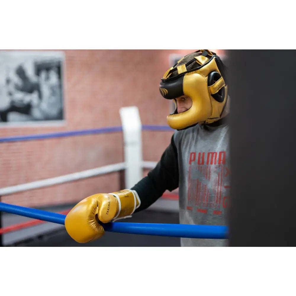 Boksehansker TIGER GOLD V, gullfarget hanske og hjelm sett fra siden mot ringtau i boksering.