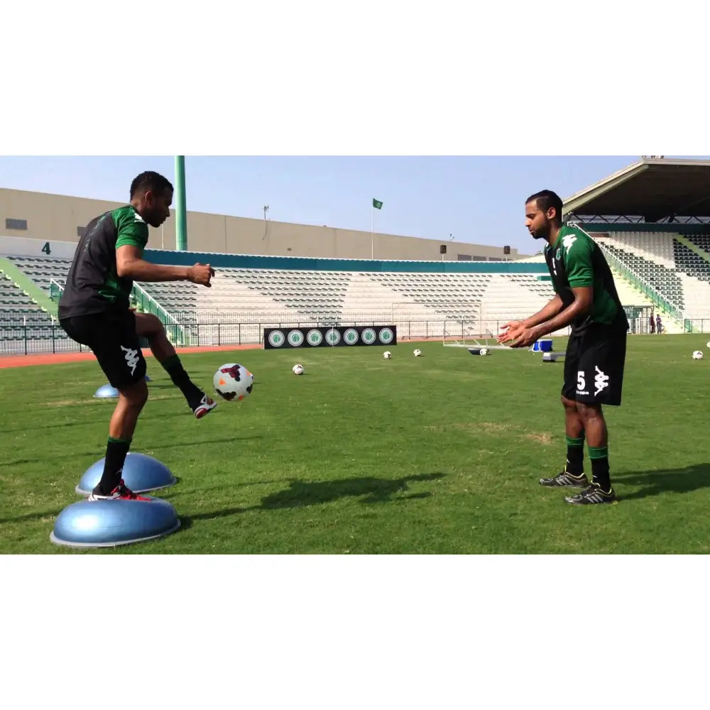 Bosu balanseputer i bruk utendørs, én mann balanserer på to blå puter mens han sparker fotball til en medspiller.