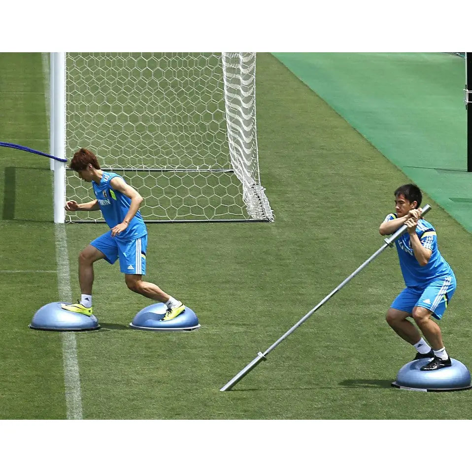 Bosu balansepute brukt utendørs, to menn trener balanse på gress med fotballmål i bakgrunnen.