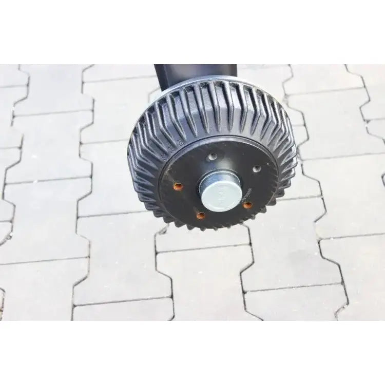 Aksling tilhenger fra AL-KO, sort riflet metall med sentral bolt, sidebilde mot grå belegningsstein.