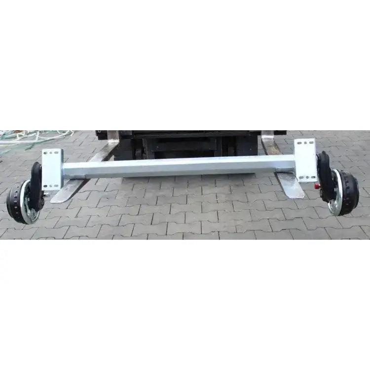 Aksling tilhenger AL-KO 1500mm, frontvisning av hel metallaksel med trommelbremser, uten hjul, på brostein.