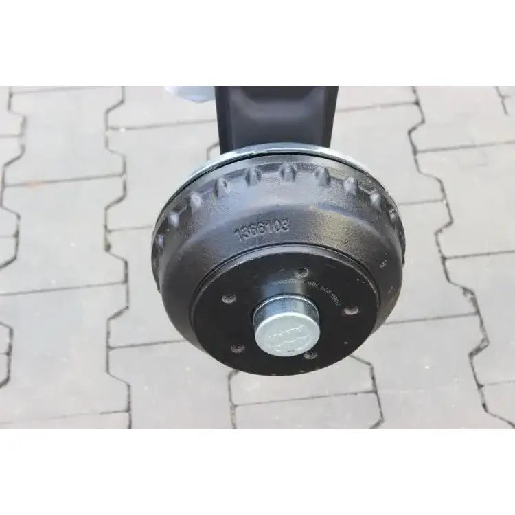 Aksling til tilhenger, sort sylinderformet metall med sentral bolt og preget nummerserie, nærbilde mot belegningsstein.