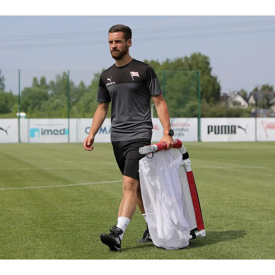 Brettport Akademiet-mål bæres av mann i svart treningstøy på fotballbane i dagslys.