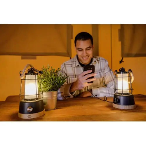 Campinglampe CAL 1 retro lyser på trebord ved siden av plante og annen lykt, ung mann i bakgrunnen.