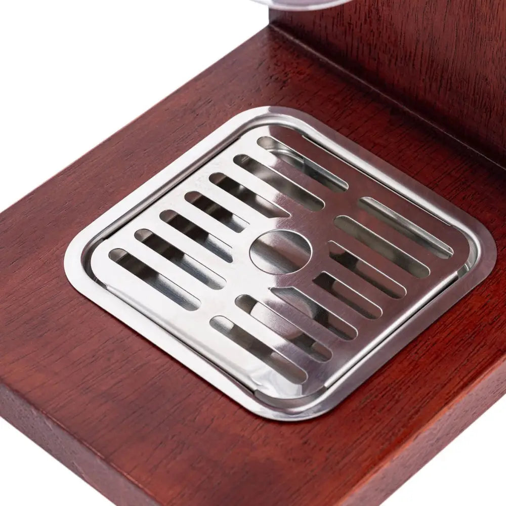 Kvadratisk dryppbrett i blankt metall, innfelt i mørk brun treplate, sett fra skrå vinkel.