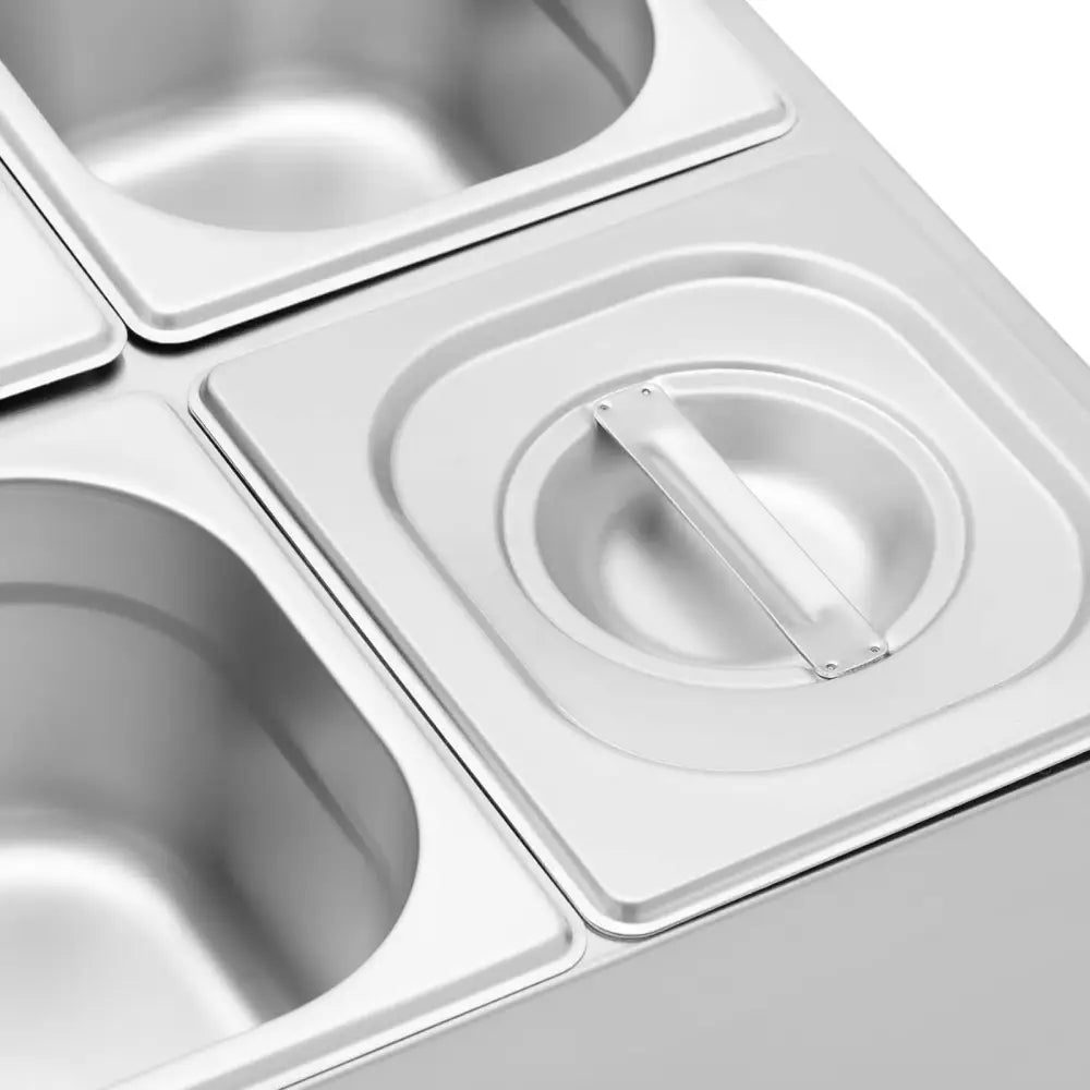 Displaystativ med fire GN 1/6 beholdere i matt stål, én med lokk og håndtak, sett skrått ovenfra.