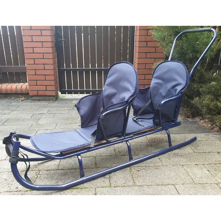 Dobbel tandem akebrett i blått, sidevisning med to polstrede seter, armlener og metallskyvestang på brostein.