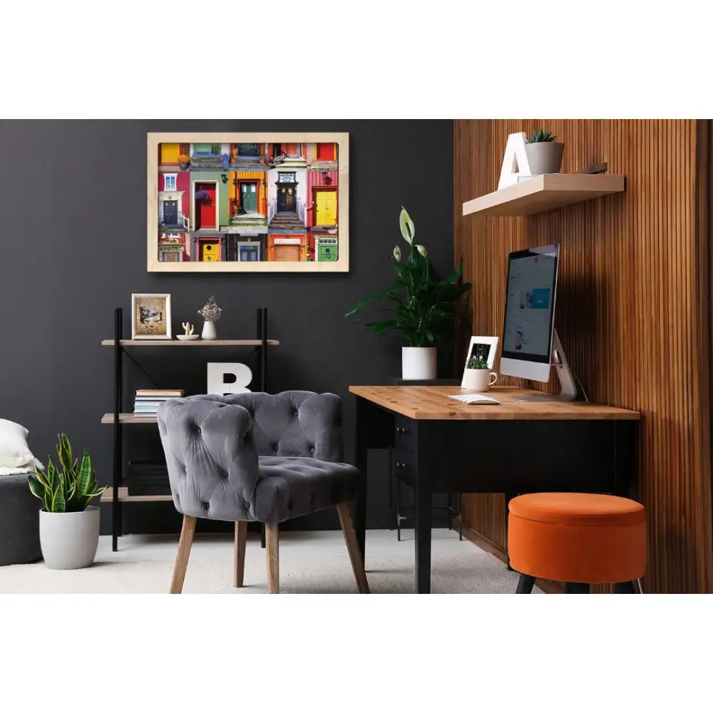 Bilde med fargerike dører henger på mørk vegg over trepult med dataskjerm, oransje puff og grå lenestol.