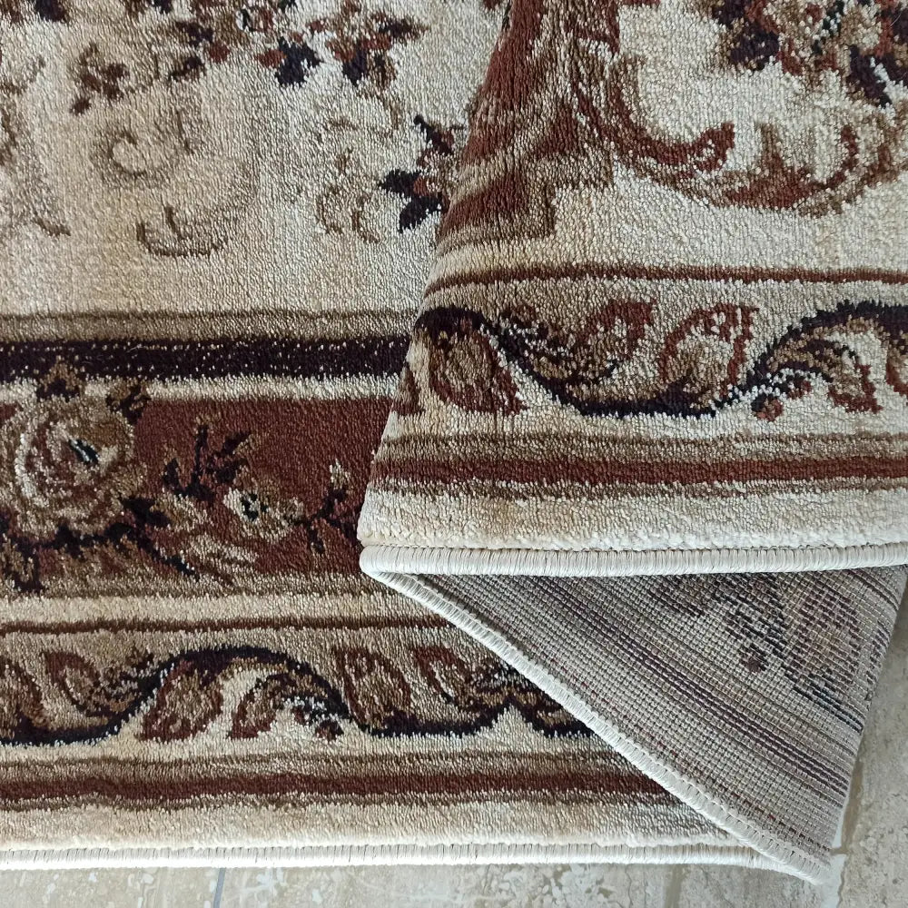 Eksklusivt teppe i beige og brunt, floralt mønster, dekorativ kant og oppbrettet hjørne som viser baksiden.