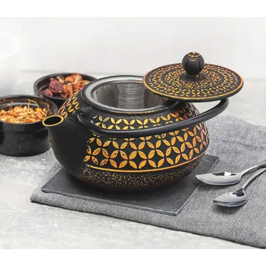 Tekanne i støpejern med gullfarget blomstermønster, lokket litt åpent og infuser synlig, på grå matte.