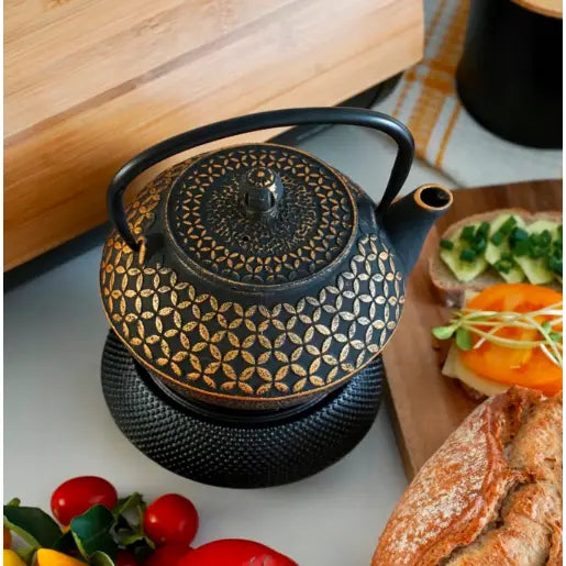 Elegant tekanne i sort støpejern med gullmønster, sett ovenfra på kjøkkenbord med matvarer og underlag.