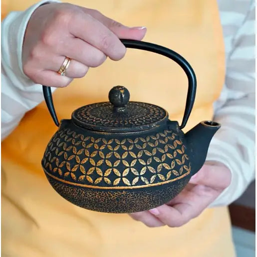 Elegant tekanne i støpejern med gyllen dekor, holdes i to hender, sett fra siden under servering.