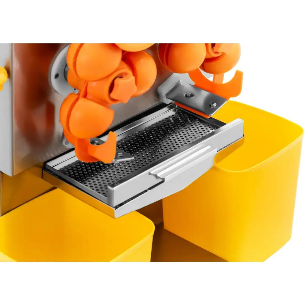 Elektrisk appelsinpress, nærbilde av oransje plastkomponenter og perforert metallrist over gule beholdere.