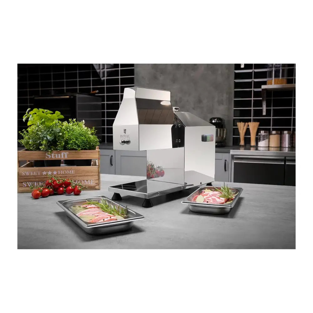 Elektrisk kjøttkvern Steaker maskin i rustfritt stål, sidevisning med rått kjøtt, urter og tomater på kjøkkenbenk.