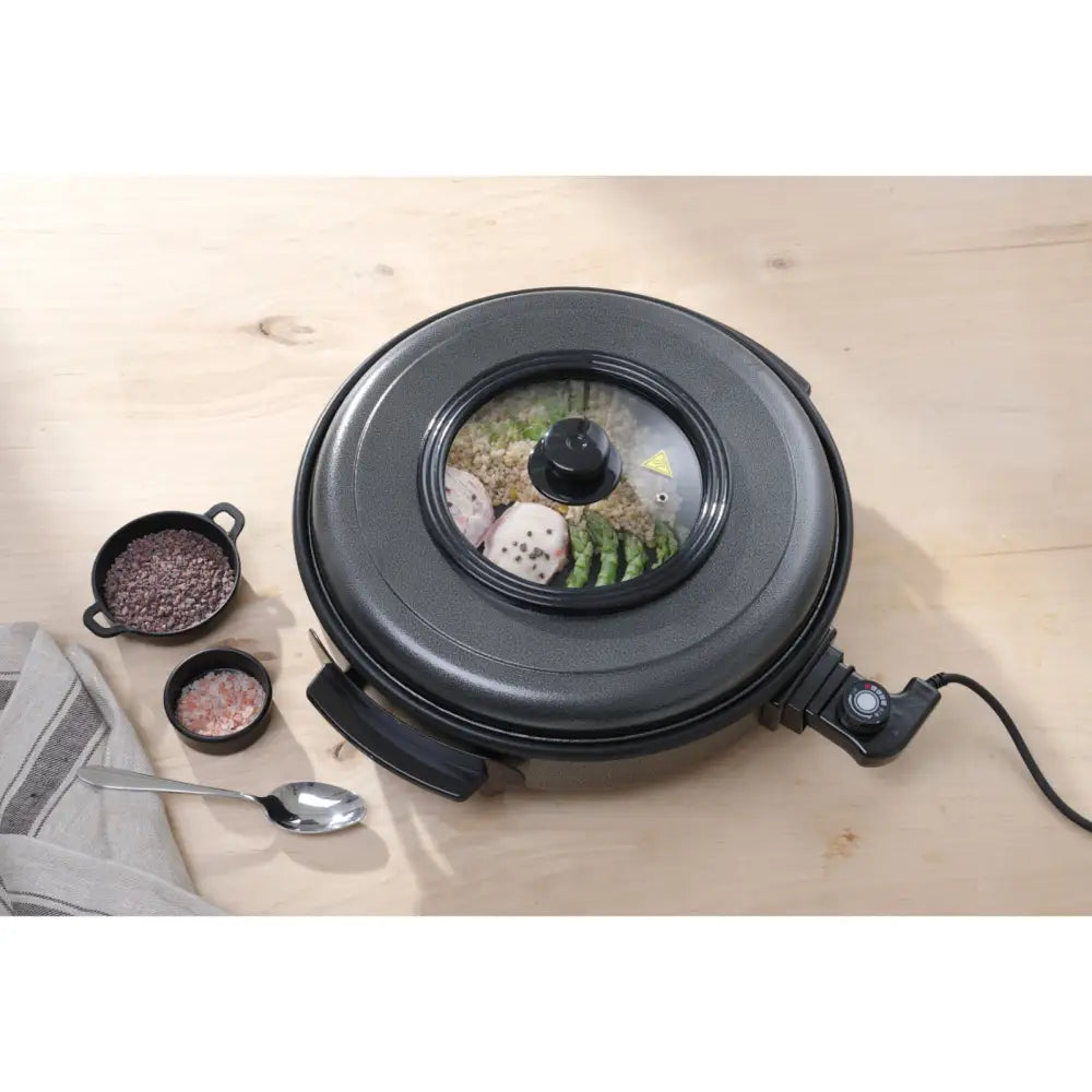 Elektrisk stekepanne i sort metall med gjennomsiktig lokk, mat synlig inni, skåler og skje ved siden av.