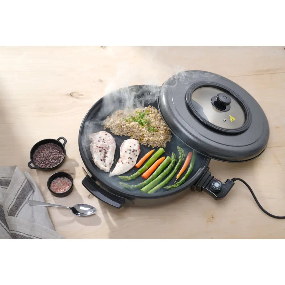 Elektrisk stekepanne i sort med glasslokk, fylt med dampende kyllingfilet, grønnsaker og ris, sett ovenfra.