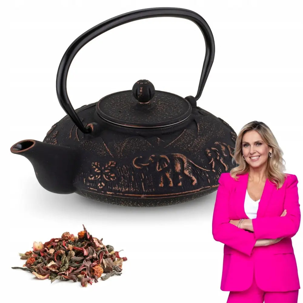 Sort emaljert støpejernstekanne med elefant- og blomsterrelieffer, løs te foran og kvinne i rosa dress ved siden av.