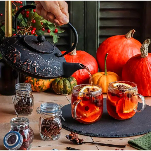 Sort emaljert støpejern tekanne med mønster, heller varm drikke i glasskrus med appelsin, krydderglass og gresskar bak.