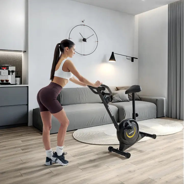 Ergometersykkel Zipro One, sort modell med gule detaljer i bruk av kvinne i lys, moderne stue med sofa.