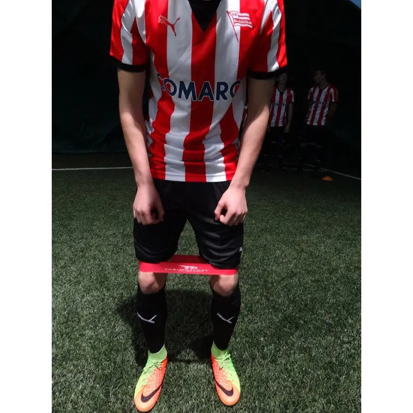 Fitnessgummi, rødt treningsbånd med medium motstand brukt rundt lårene på kunstgress under fotballtrening.