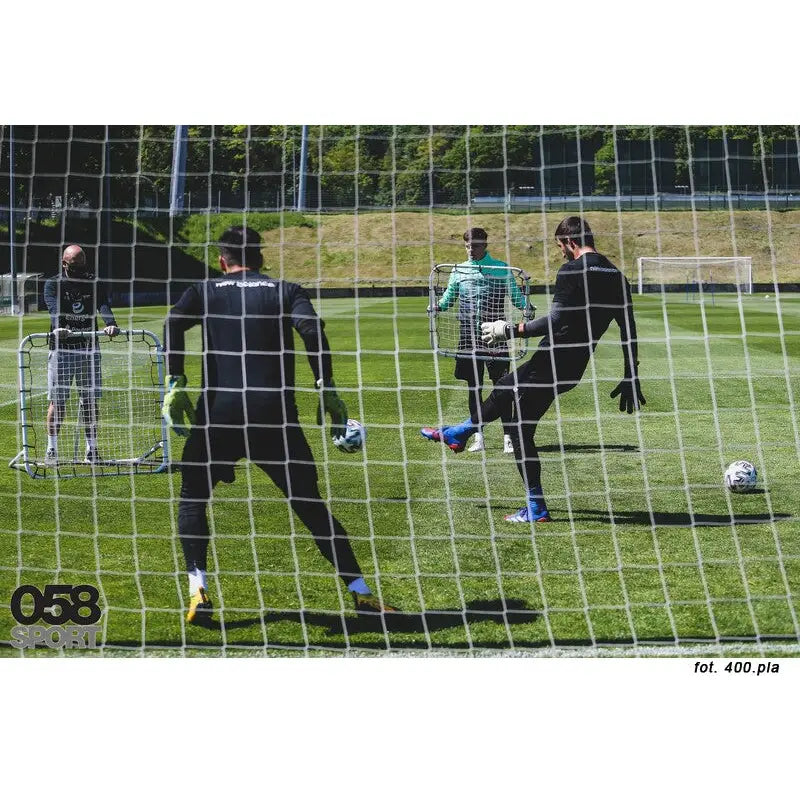 Fjærbrett ramme med nett, fire personer trener fotball med reboundere foran fotballmål på gressbane.