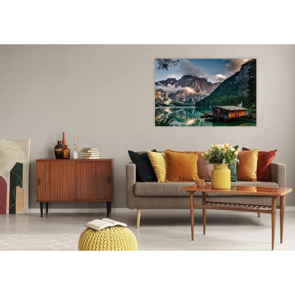 Fjell, hytte og innsjø lerretsbilde over lys grå sofa med fargerike puter og lavt trebord i stue.