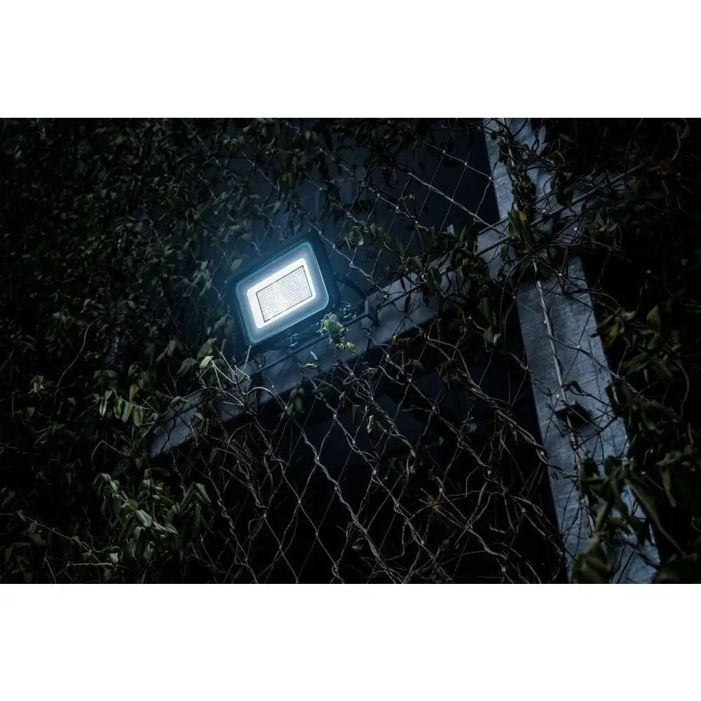 LED flomlys JARO, montert på gjerde blant grønne planter, lyser opp uteområde i mørket fra lav vinkel.