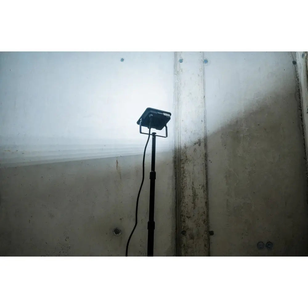 Flomlys LED JARO på stativ sett fra siden, lyser mot betongvegg med synlig strømkabel.