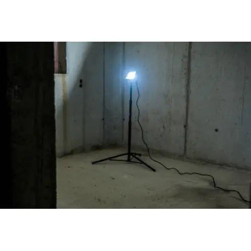 Flomlys på stativ LED, skråvisning i mørkt betongrom med tydelig lysstråle og synlig strømledning på gulvet.