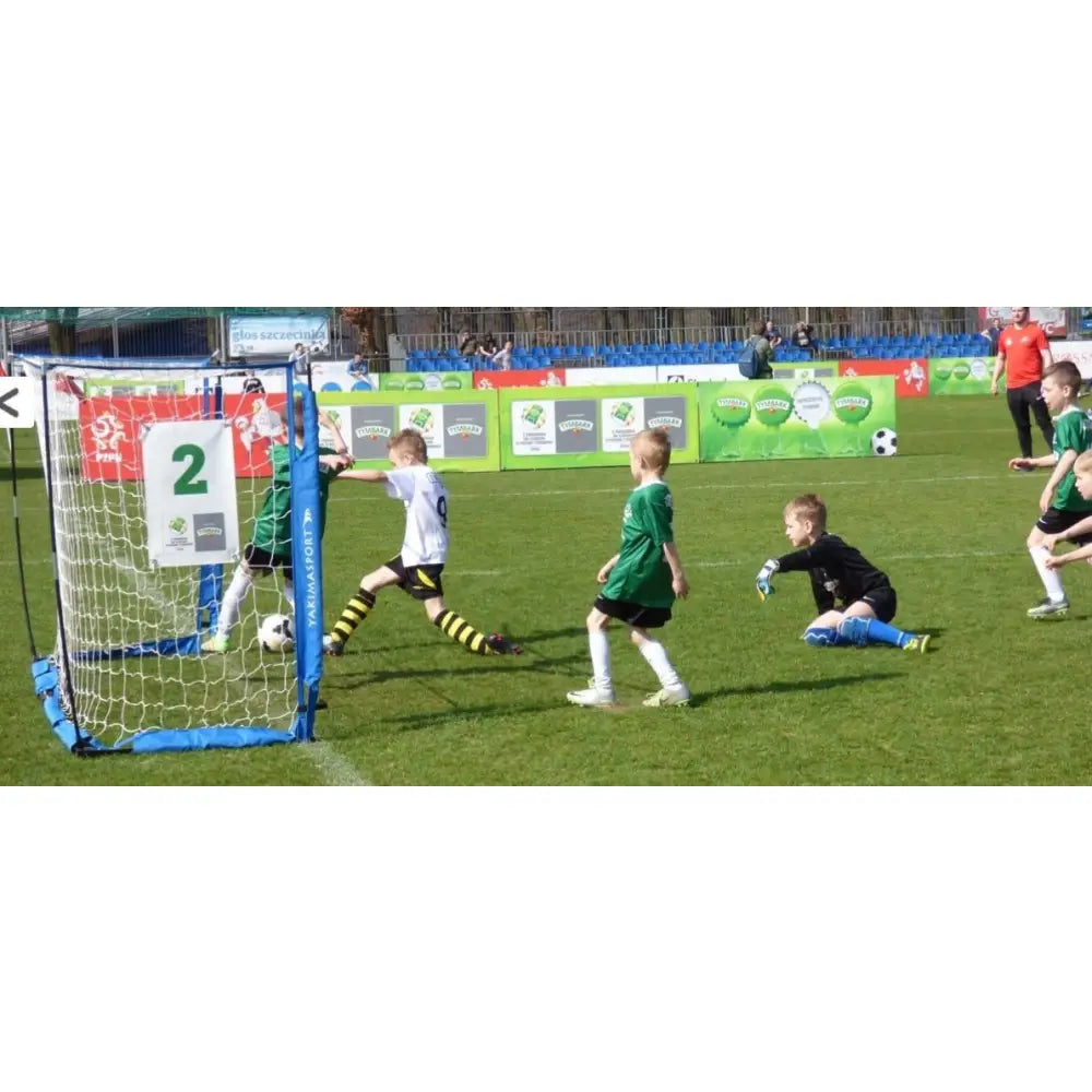 Foldbart blått fotballmål UNI ŻAK, nærbilde av målområdet mens barn spiller kamp på gress.